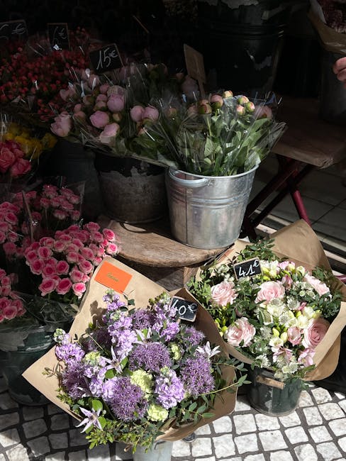 A display of fresh floral arrangements at a flower market, featuring bouquets with pink roses, purple hydrangeas, and white roses, wrapped in brown paper and placed in metal buckets and plastic vases 