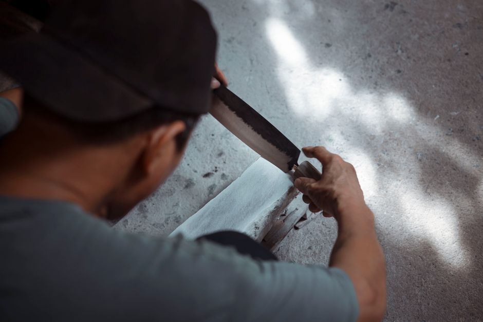 A craftsman wearing a dark cap and a grey shirt is seen from behind, using a large handsaw to cut a piece of white material, possibly paper or fabric, on a textured surface. The setting appears to be 