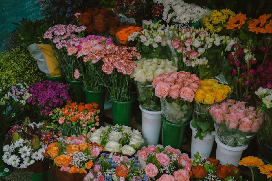A vibrant display of fresh flowers arranged in white and green buckets, featuring a variety of roses in soft pink, peach, and yellow hues, along with clusters of white, purple, and orange blooms, as w