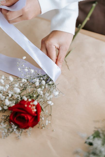 Close-up of a person’s hands, with one hand holding a white satin ribbon and the other steadying a bouquet of fresh flowers on a wooden surface. The bouquet includes a vibrant red rose, white baby's