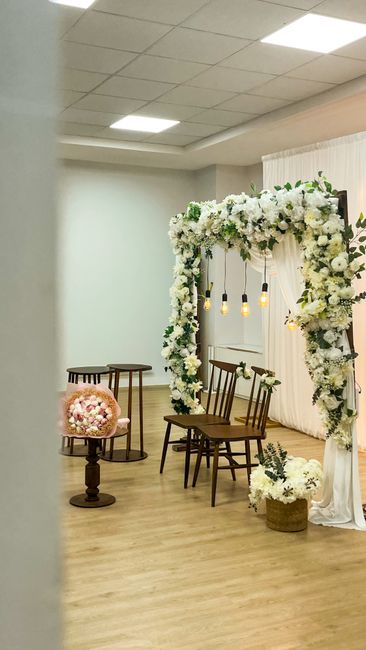 A decorative wedding floral arrangement featuring a large square arch adorned with an abundant display of white flowers, including roses, hydrangeas, and jasmine, interwoven with greenery. The arch is