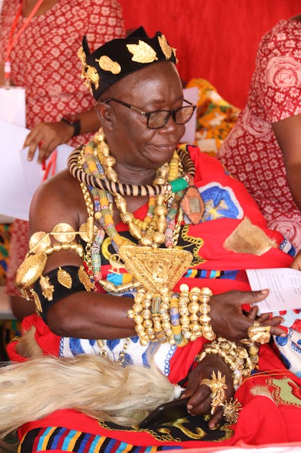 A woman dressed in traditional vibrant Maasai attire, adorned with numerous gold and colorful beaded jewelry including necklaces, bracelets, and earrings. She is seated on a red patterned fabric, hold